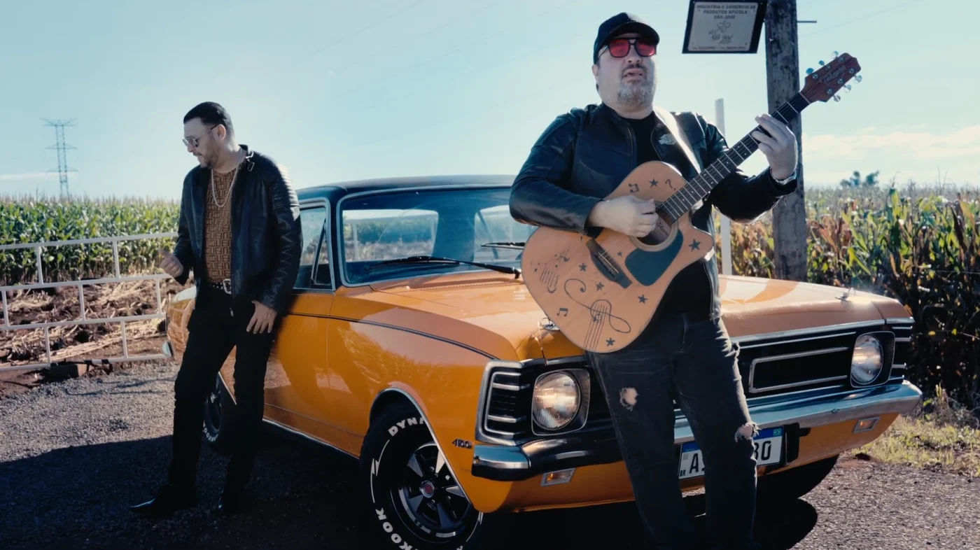 Músicos com violão ao lado de um carro antigo em cena de videoclipe